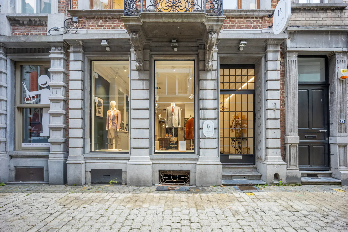 Storefront of a tailor shop with mannequins in the windows, stone facade, and cobblestone street.
