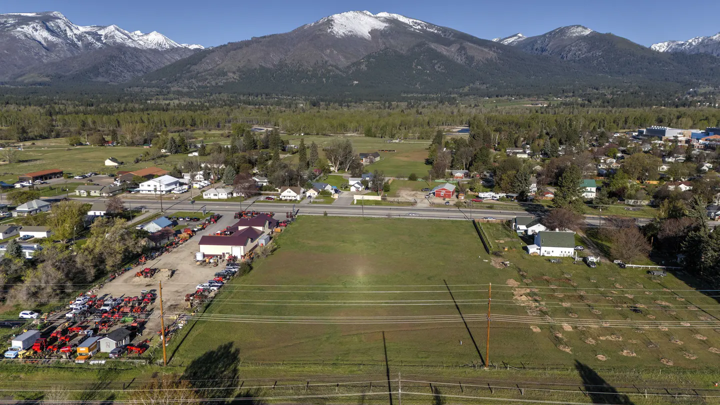 Aerial view of a rural town with a large green field, surrounded by houses, trees, and snow-capped mountains in the background.