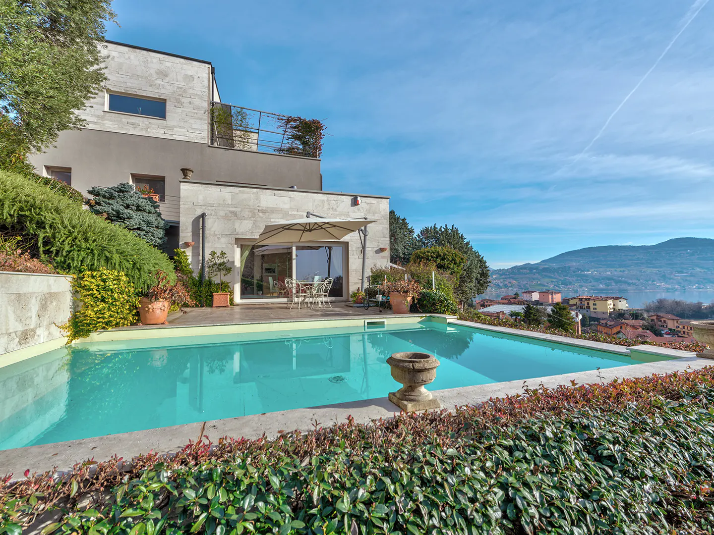 A modern home with a turquoise pool, stone patio, and manicured hedges on a sunny day.
