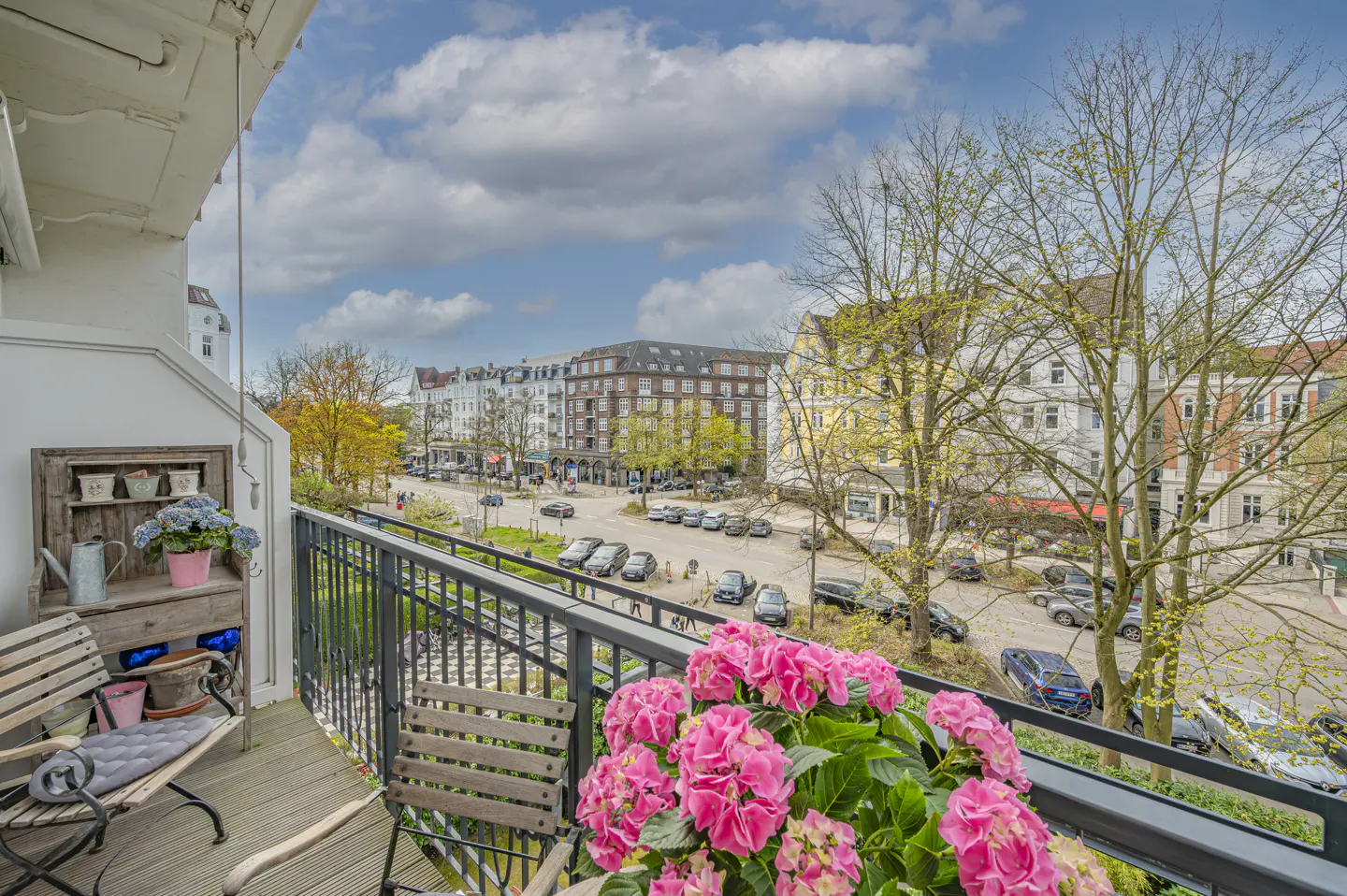 Balcony view with pink hydrangeas, wooden chairs, and a bench. City street with cars and buildings in the background under a cloudy sky.