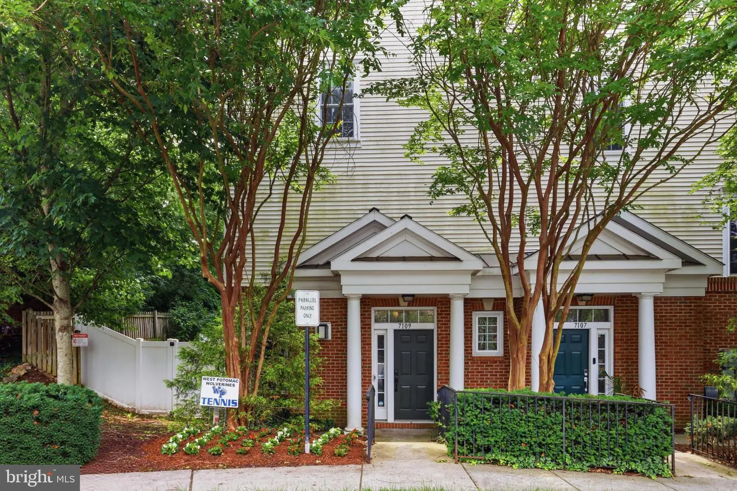 Exterior view of two brick townhomes with dark doors, white columns, and mature trees in front of a sidewalk.