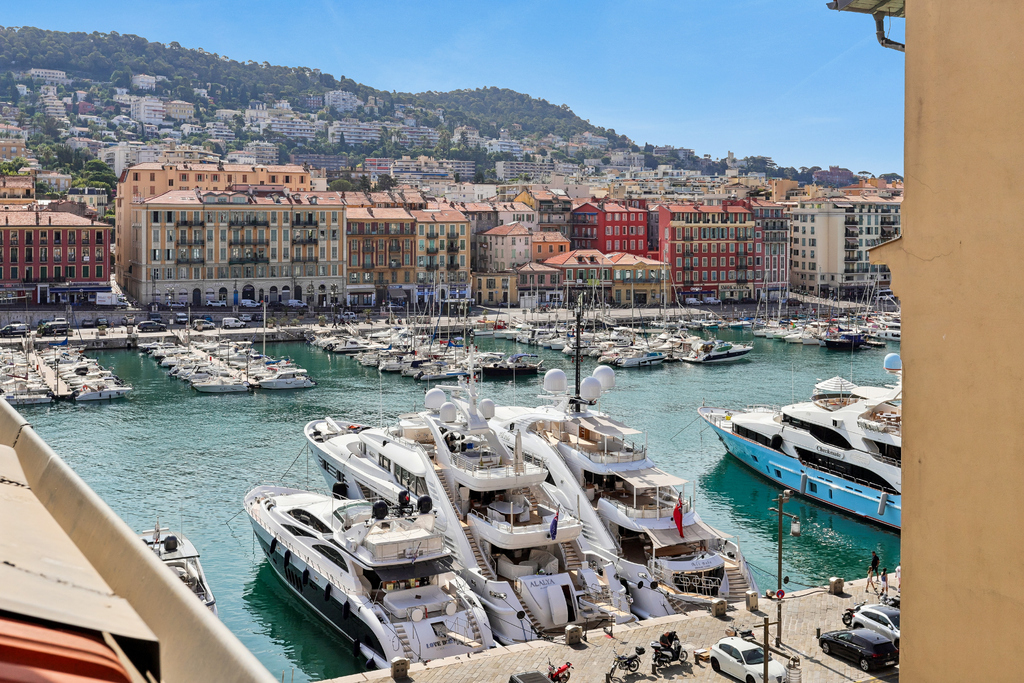 View of a harbor in Nice, France, featuring luxury yachts docked in turquoise water before a backdrop of colorful buildings. [AI-generated text]