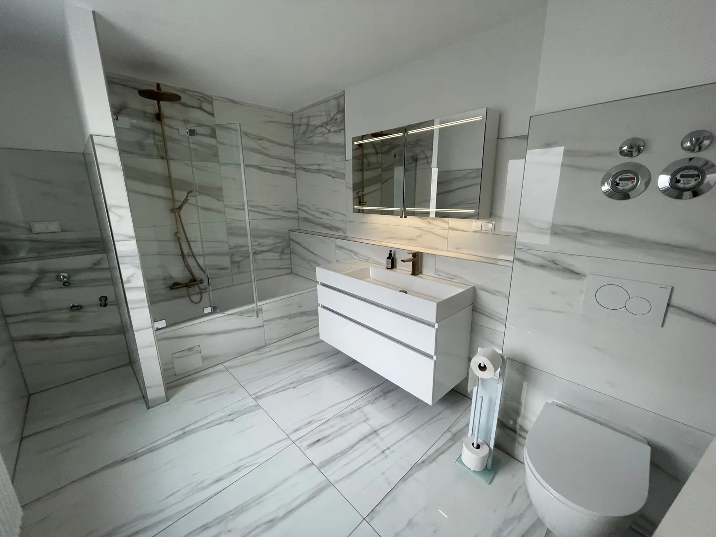 Bright bathroom with marble tile, featuring a white floating vanity, toilet, and glass-enclosed shower with bronze fixtures.