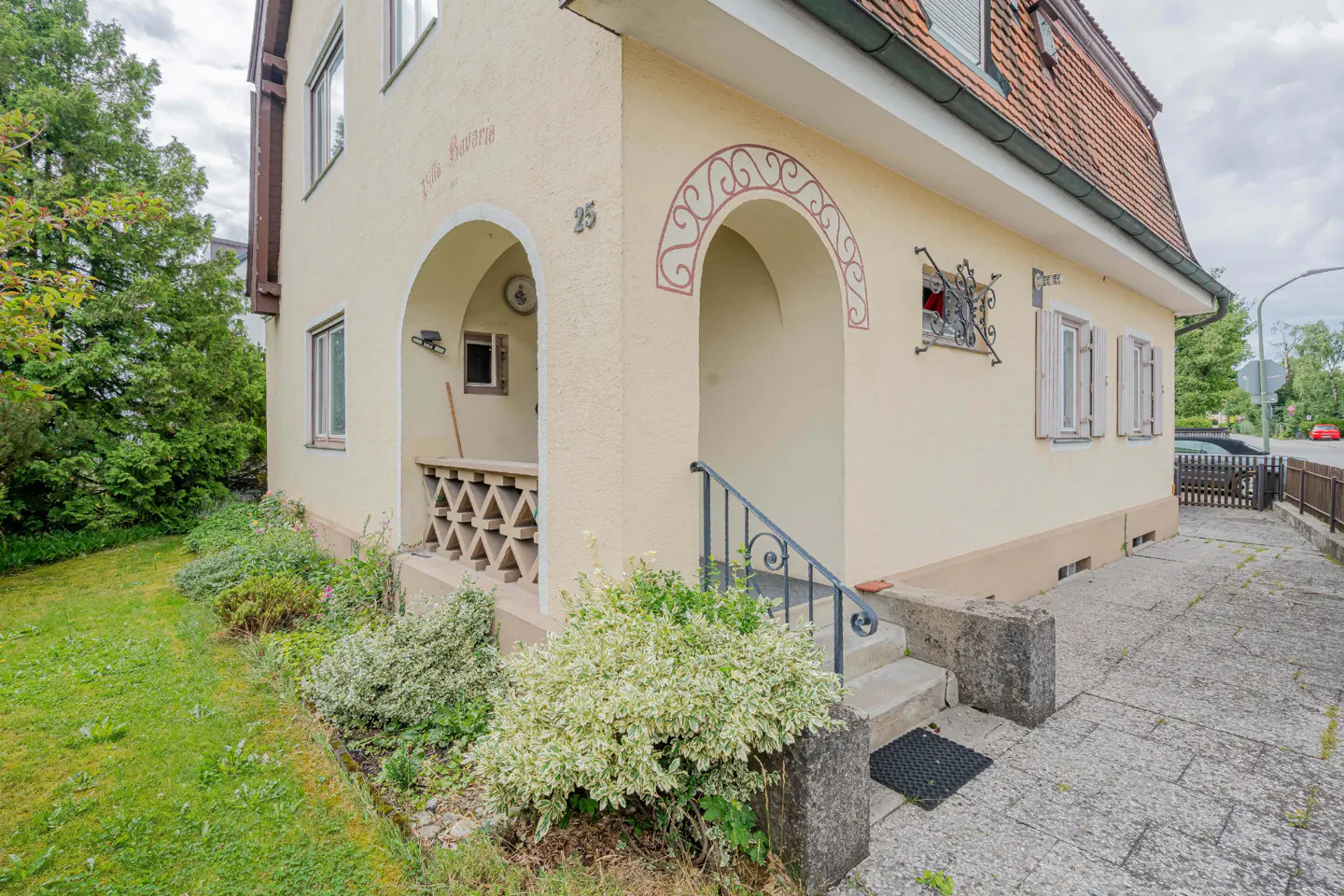 Exterior view of a two-story, light yellow house with arched entryways and a red-tiled roof. A small garden is in the foreground.
