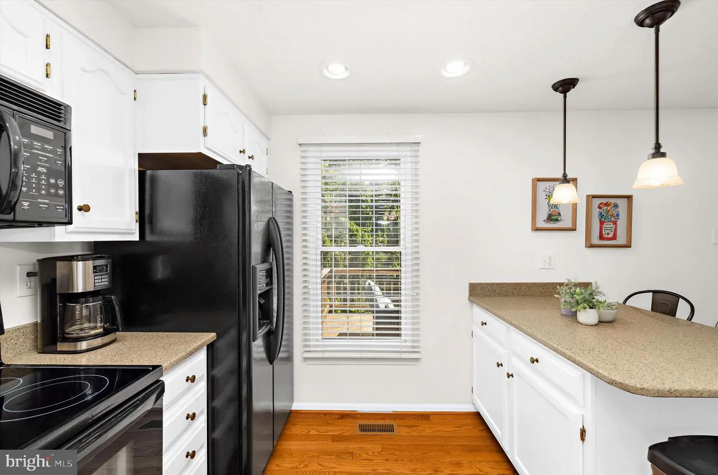 Bright kitchen with white cabinets, tan countertops, and wood floors. Black appliances include a microwave and refrigerator. A window overlooks a deck.