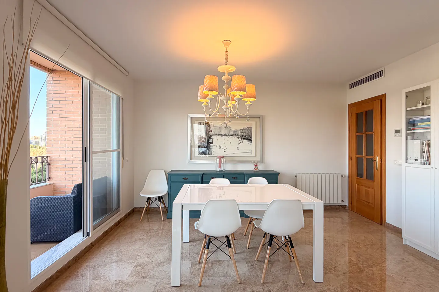 Bright dining room with white table, chairs, and marble floor. A blue cabinet sits under a framed picture. Balcony visible through sliding glass doors.