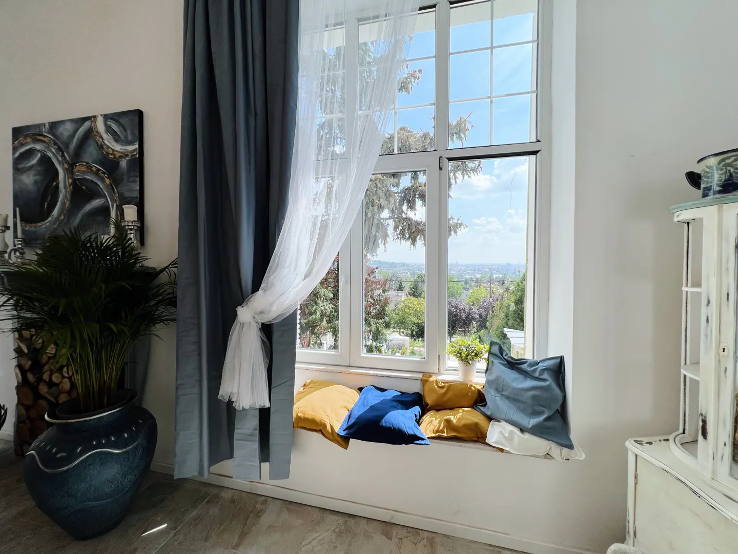 Bright room with a large window, blue and gold pillows on the sill, and sheer white curtains tied back. A potted plant sits to the left.