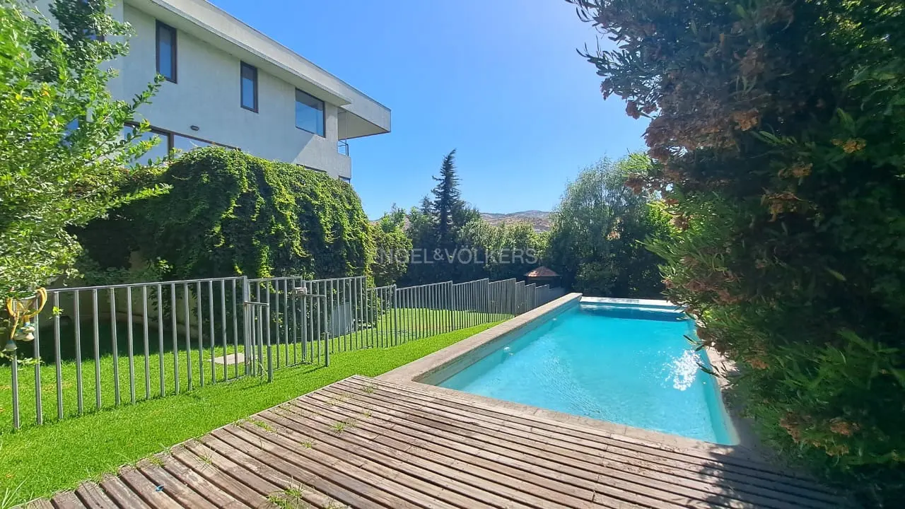 A backyard with a pool, wooden deck, and green lawn, surrounded by a metal fence and lush trees, under a clear blue sky.