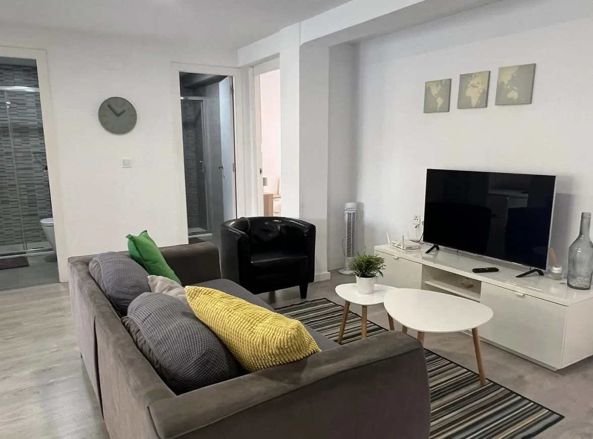 Modern living room with a grey sofa, yellow and green pillows, a black armchair, white coffee tables, and a flat-screen TV.