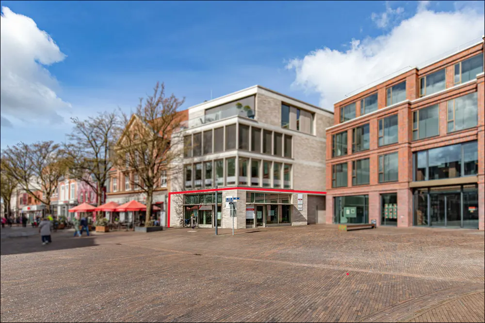 Exterior view of a modern, multi-story building with a red-trimmed storefront on a brick-paved plaza.