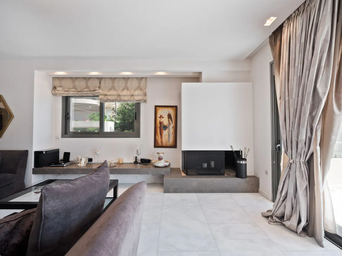 Bright living room with a modern fireplace, gray sofa, and a window with patterned blinds. Neutral-toned curtains frame a glass door.