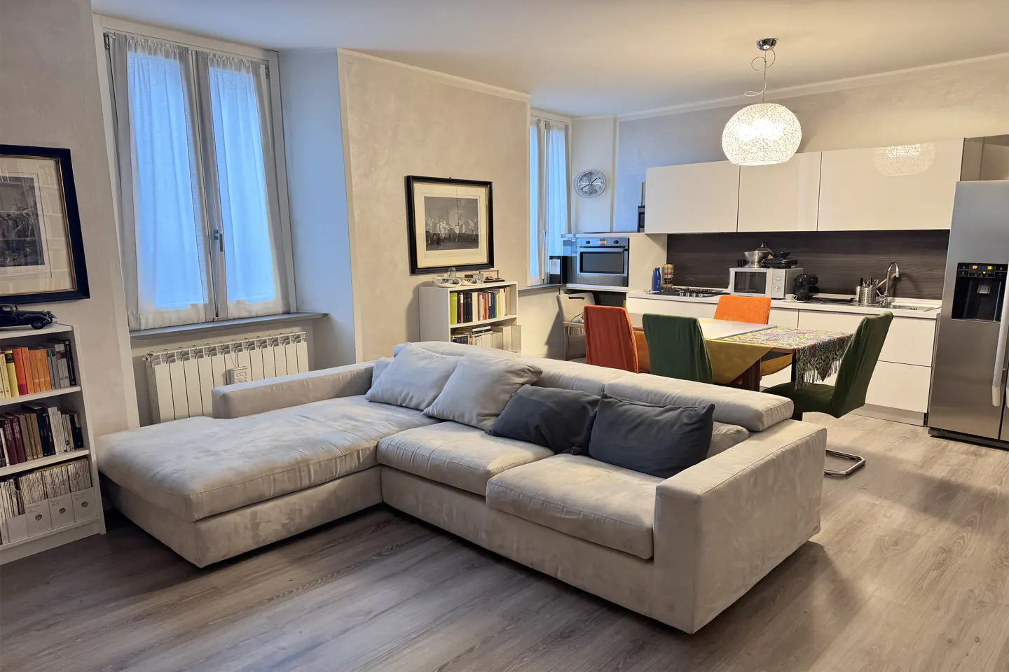 Open-concept living space with a beige sectional sofa, wood floors, and a modern white kitchen in the background.