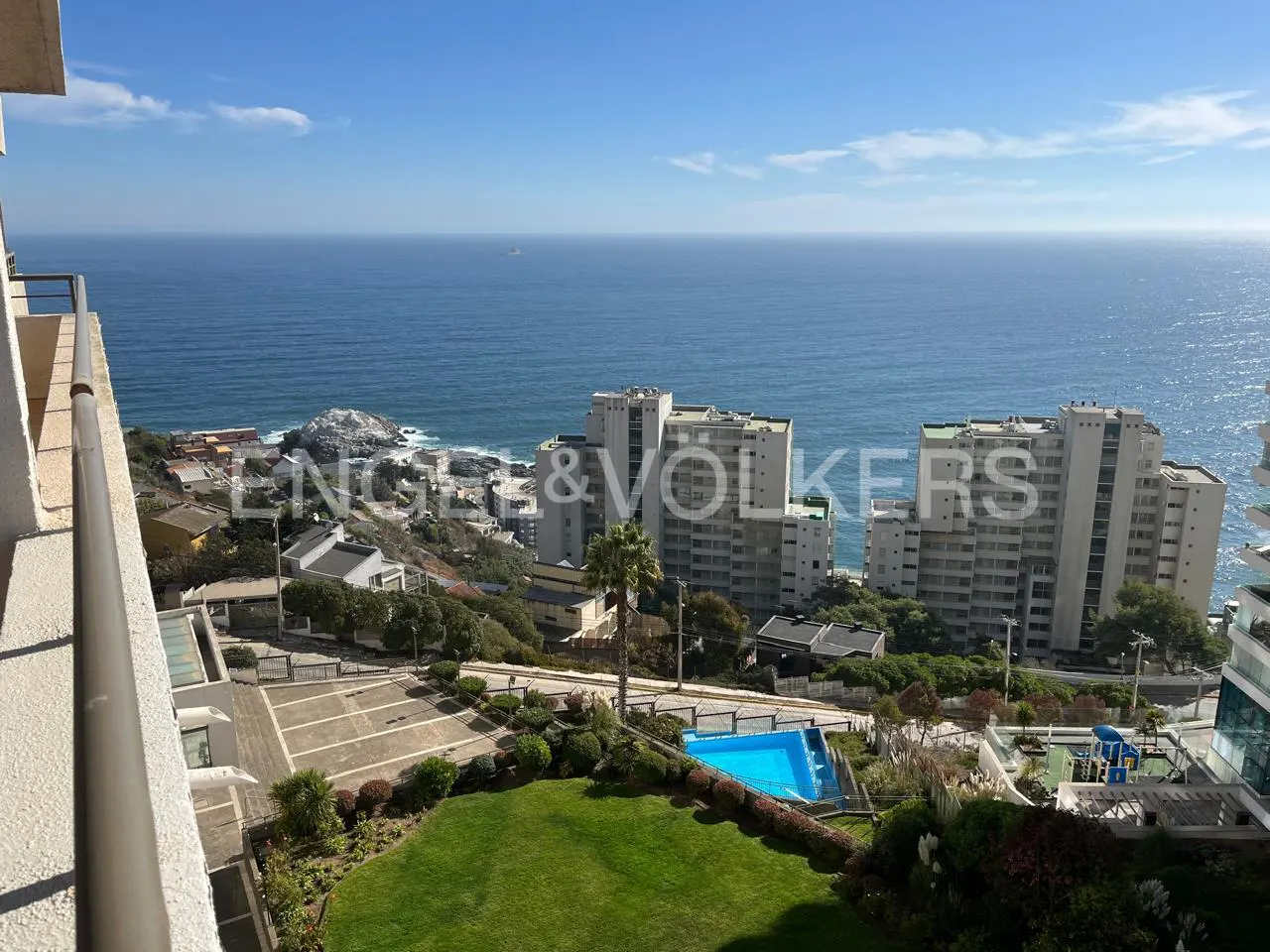 Balcony view of ocean, buildings, and pool. Blue sky above. Green lawn in foreground. Engel & Völkers logo visible.