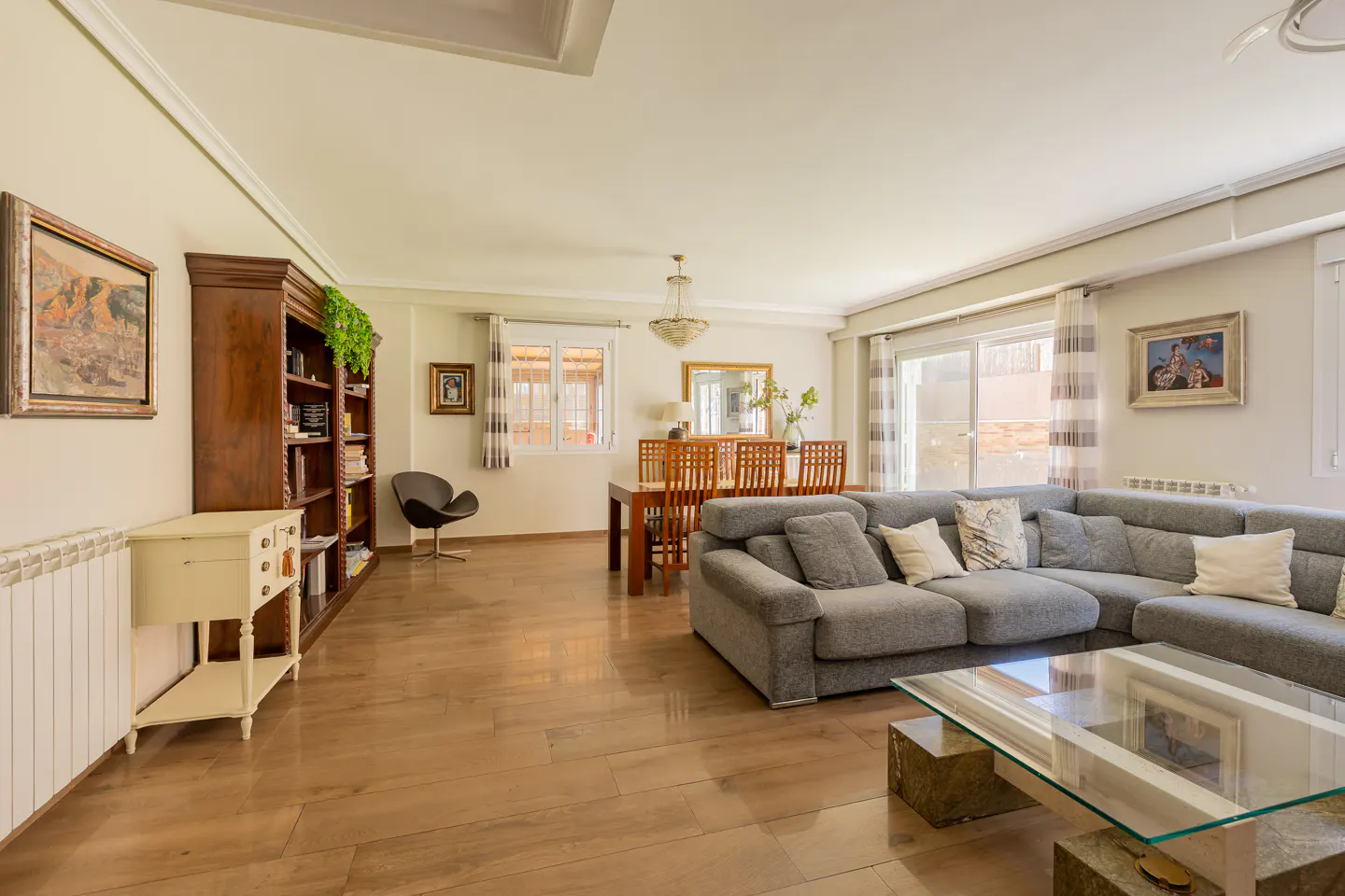 A bright, open-concept living and dining area with hardwood floors, a gray sectional sofa, and a wooden dining table.