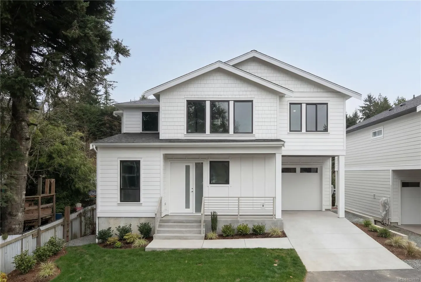 Two-story white house with black-framed windows, a front porch, and a driveway leading to a garage. Green lawn and trees surround the house.