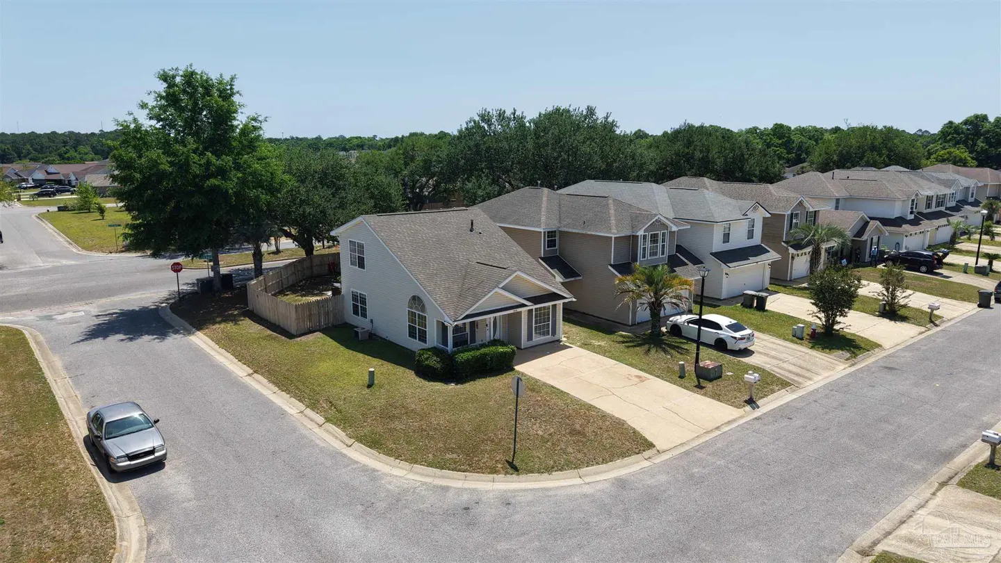 Aerial view of a suburban street with light-colored houses, green trees, and parked cars under a clear blue sky.