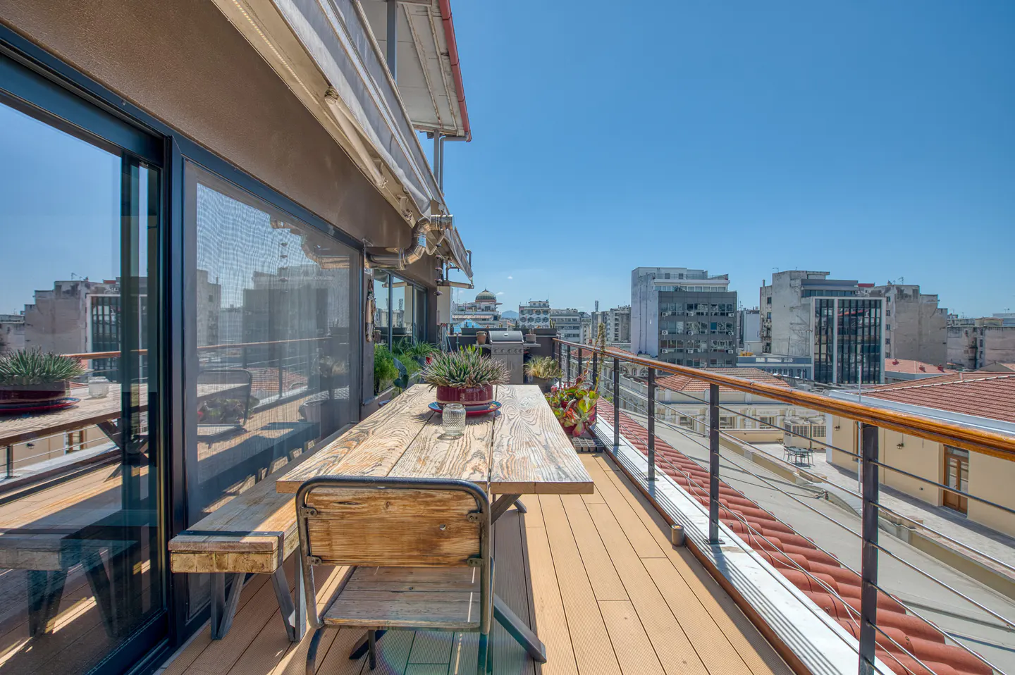 Balcony view with wooden table, chair, and bench overlooking a cityscape under a clear blue sky. Railing with wood trim and metal bars.