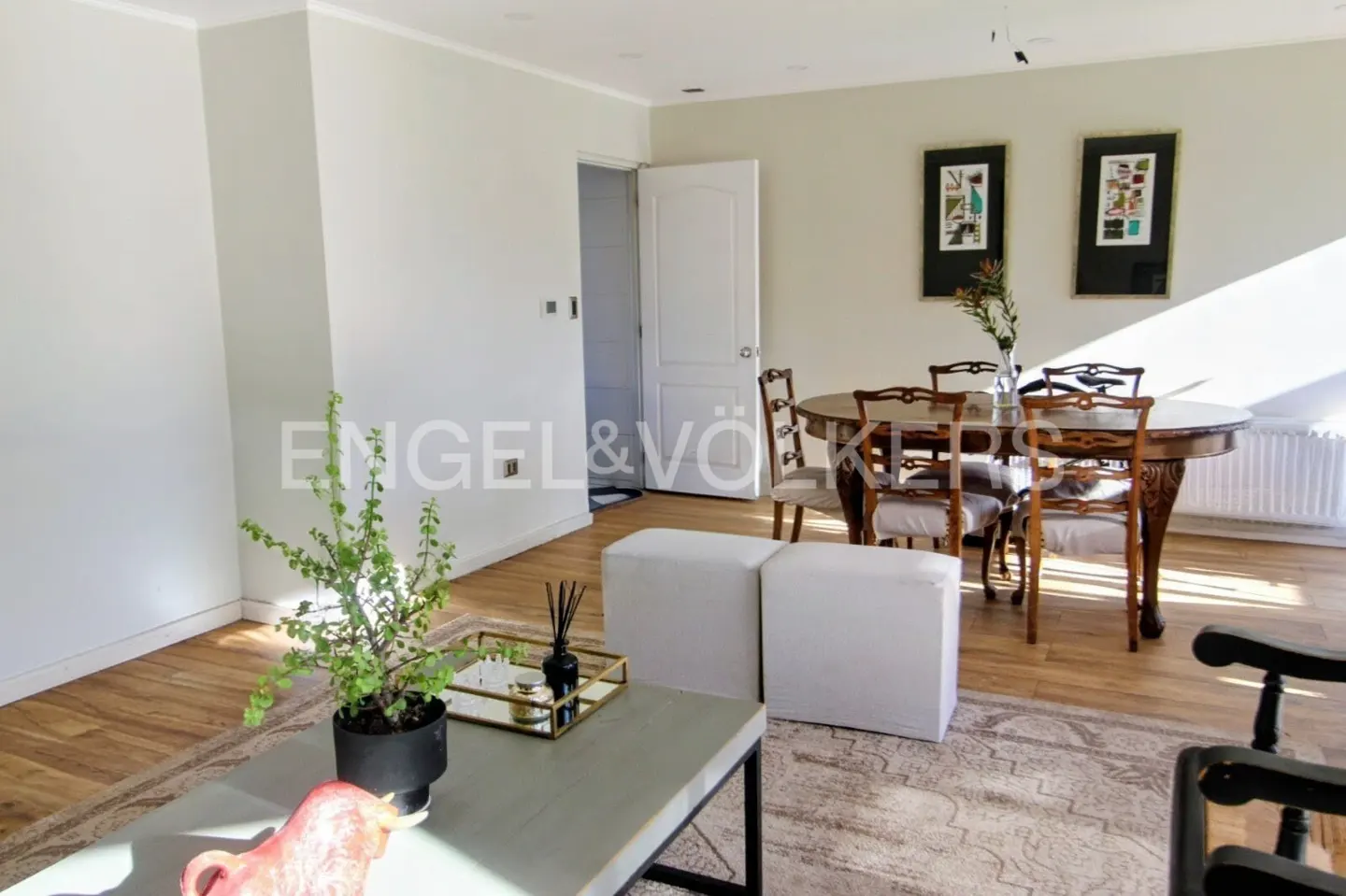Bright living room with wood floors, a dining table with chairs, and a coffee table with a plant. Two white ottomans sit on a patterned rug.