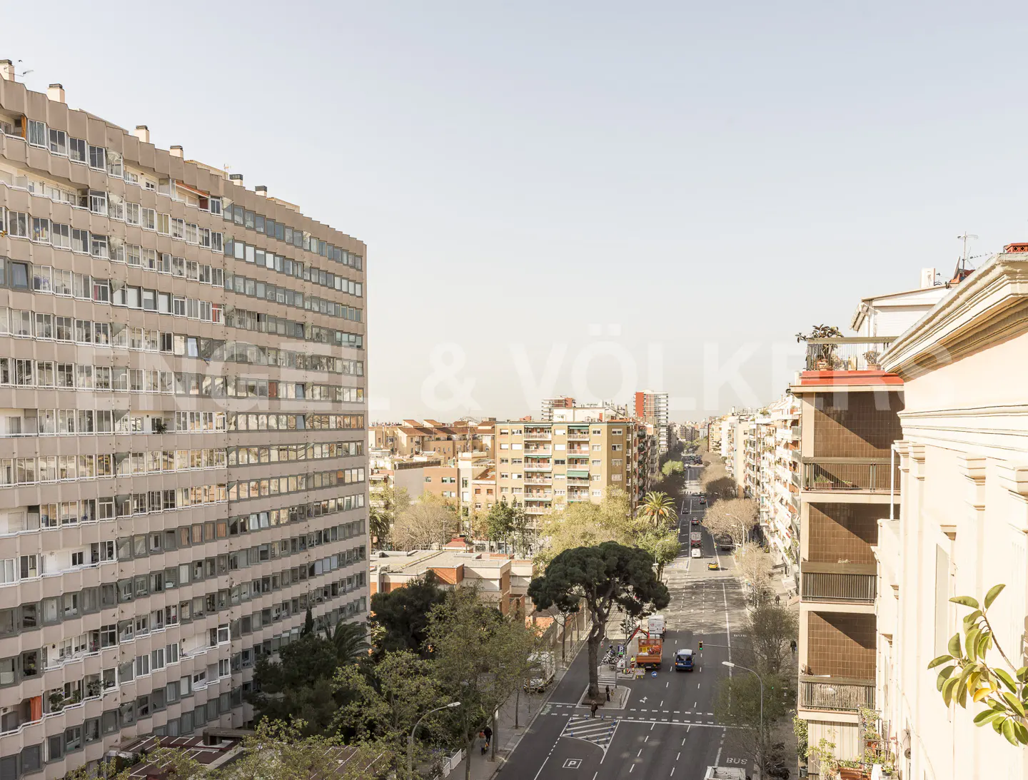 Cityscape view from a high vantage point, featuring a wide street lined with buildings and trees under a bright sky.