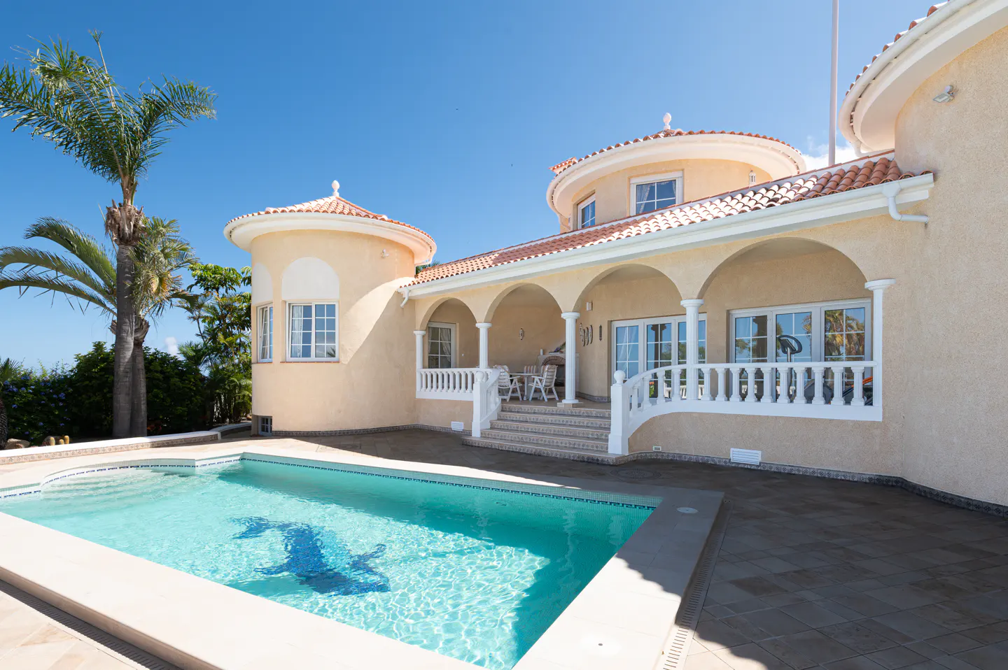 Exterior view of a tan house with a pool, arches, white railings, and a palm tree against a blue sky.