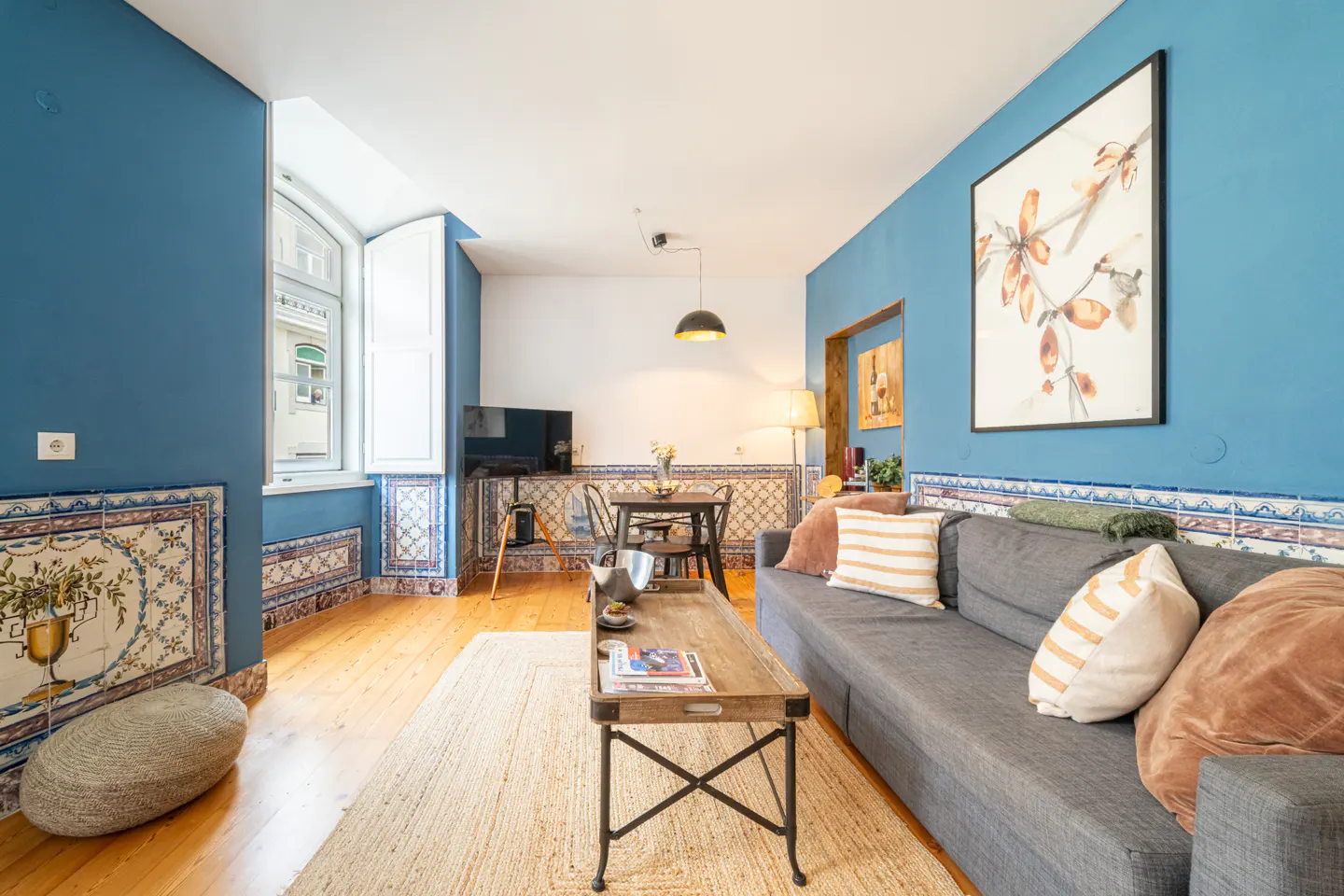 Bright living room with blue walls, tile trim, and wood floors. A gray sofa faces a wooden coffee table and dining area. A large floral print hangs on the wall.