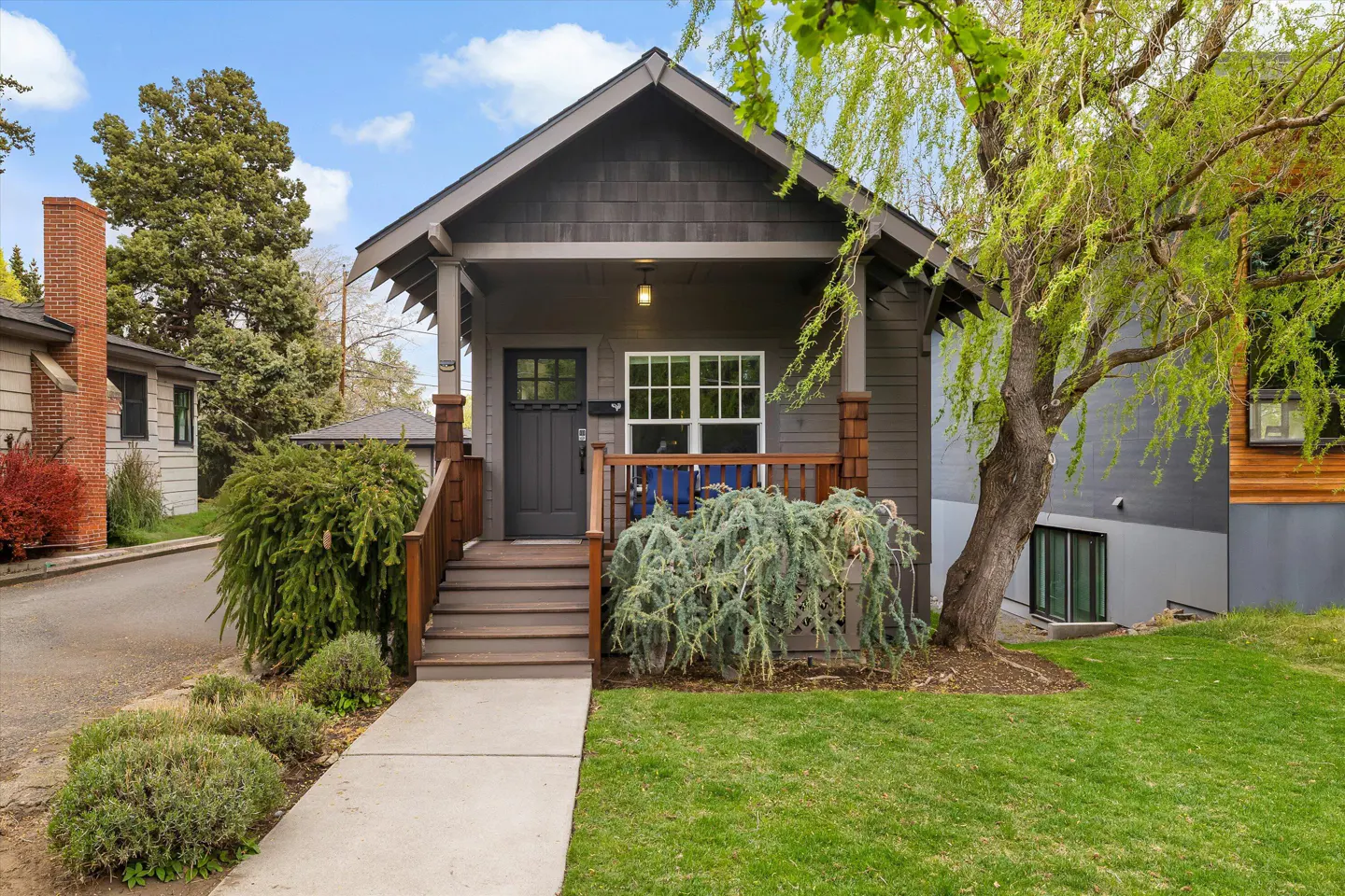 A gray, one-story house with a porch, brown trim, and a green lawn. A tree is on the right.
