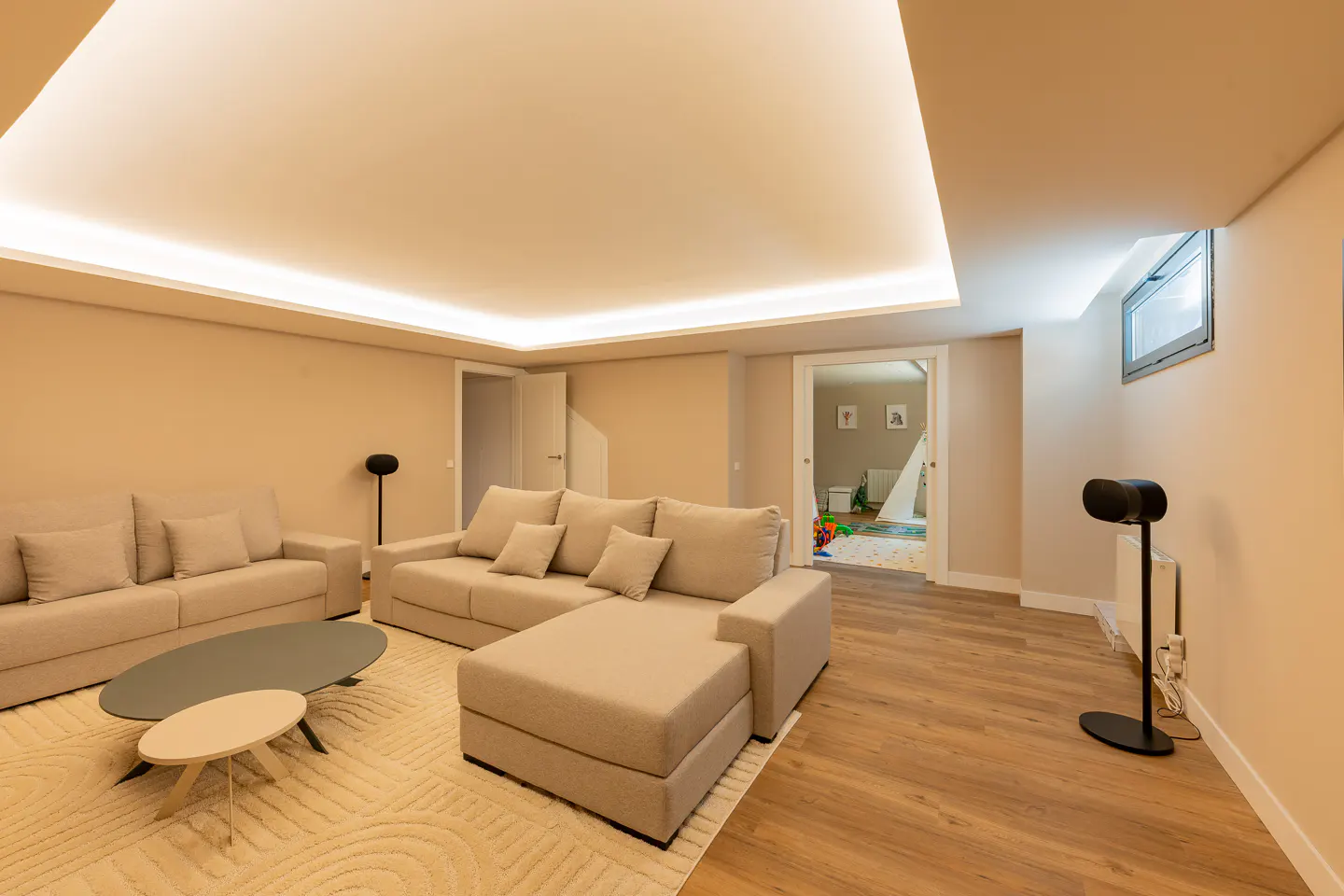 Living room with beige sofas, wood floors, and a cream rug. A doorway leads to a playroom with a teepee tent.