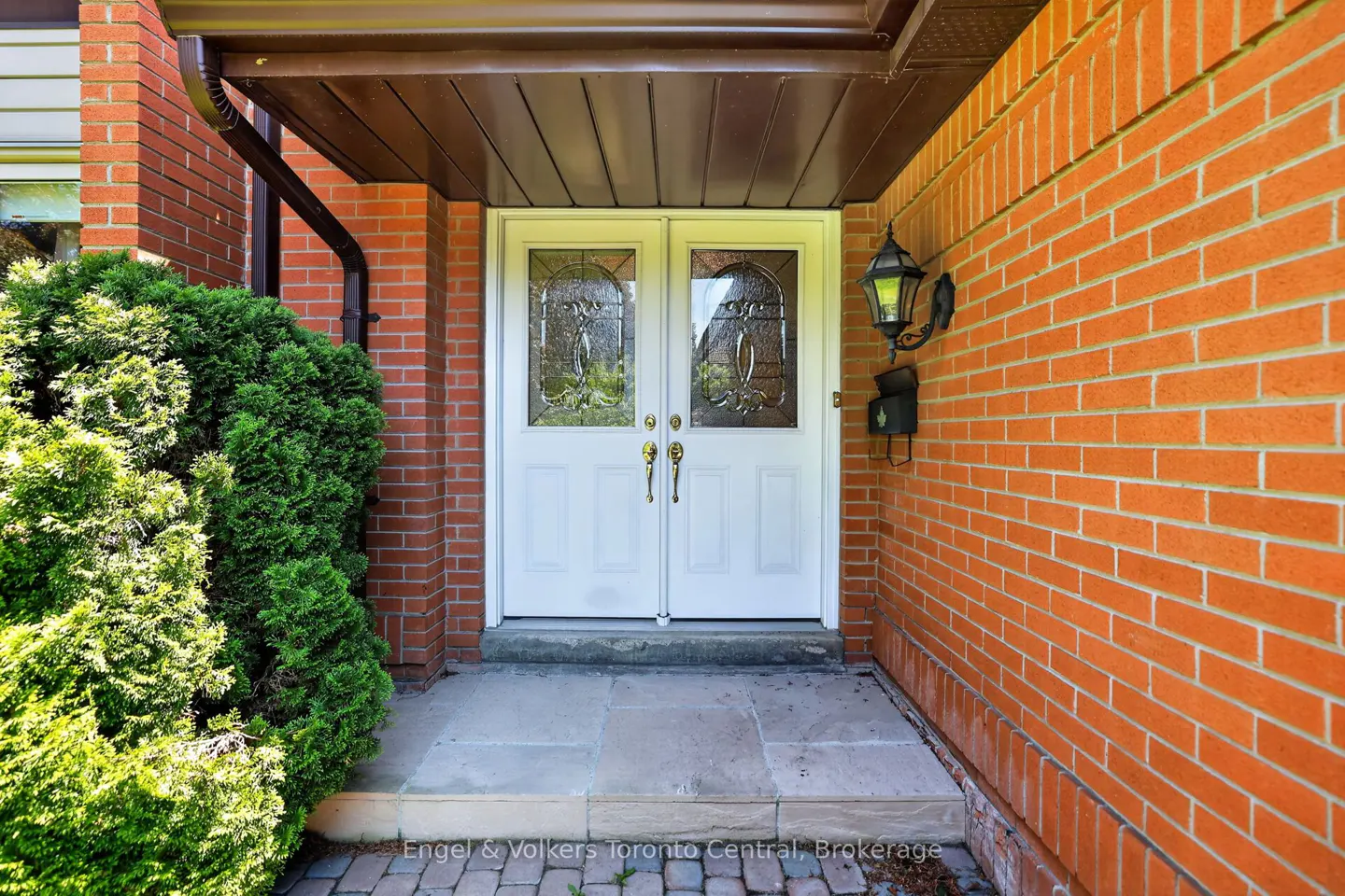 White double front doors with glass inserts set in a red brick house entrance with a stone porch and green shrub.