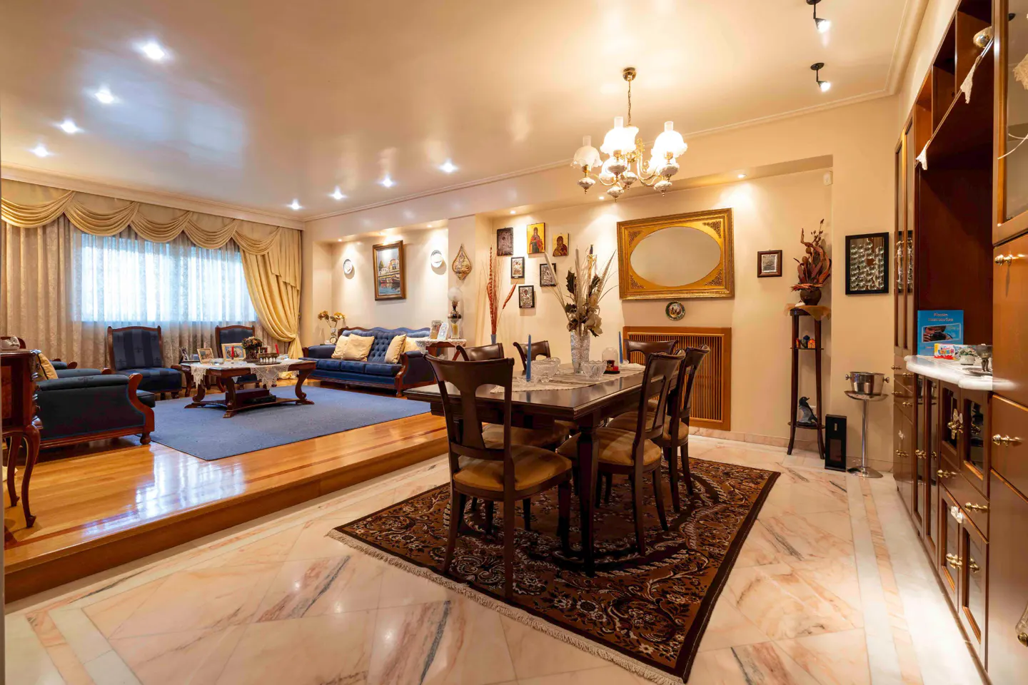 Bright living room with blue sofas on a raised platform, a dining table with chairs, and a large wooden cabinet.