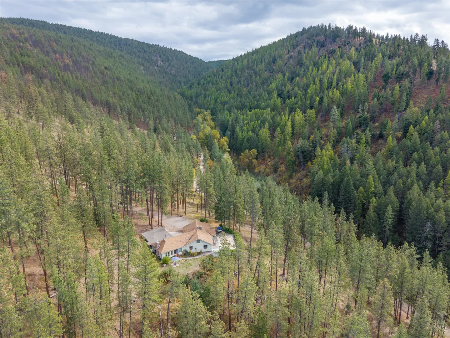 Aerial view of a light-colored house nestled in a dense forest between two hills under a cloudy sky.