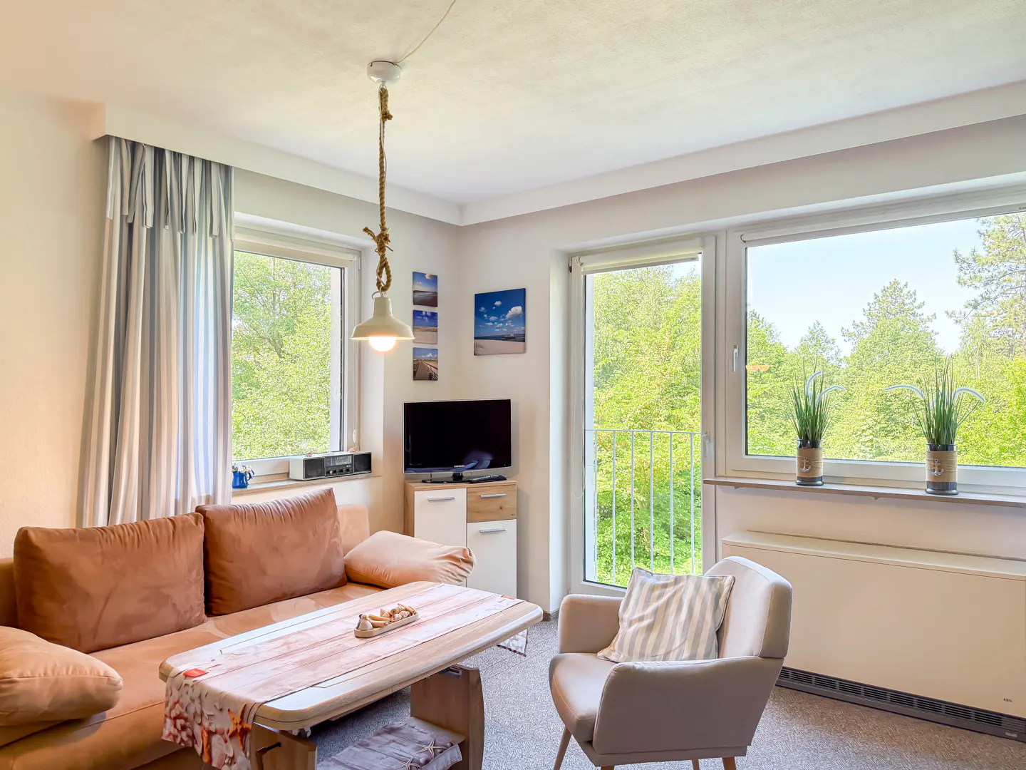 Bright living room with a tan sofa, armchair, wooden coffee table, TV, and large windows overlooking lush green trees.
