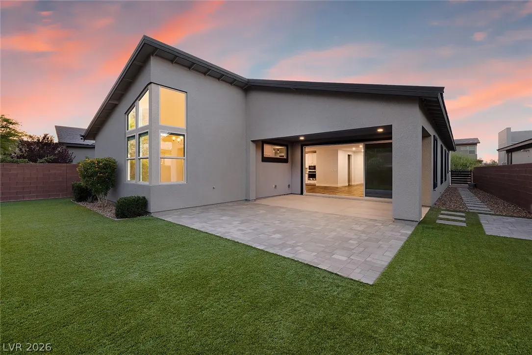 Backyard view of a gray house with a patio, green lawn, and large sliding glass doors open to the interior. The sky is pink and orange.