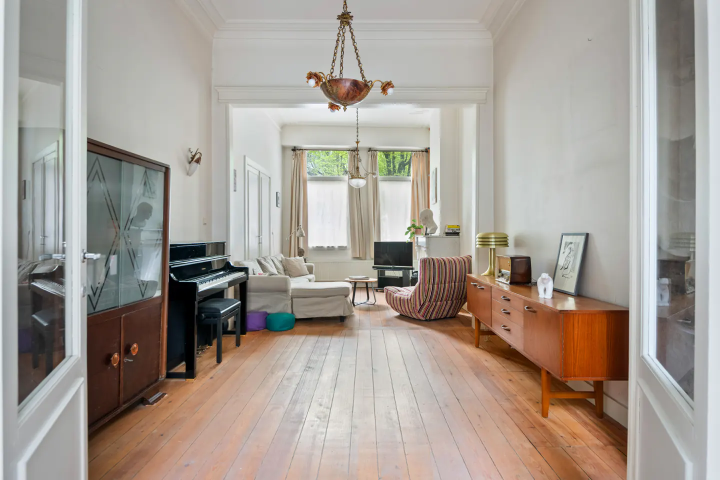 A bright living room with wood floors, a piano, a cabinet, and a striped chair. A sofa is visible in the background.