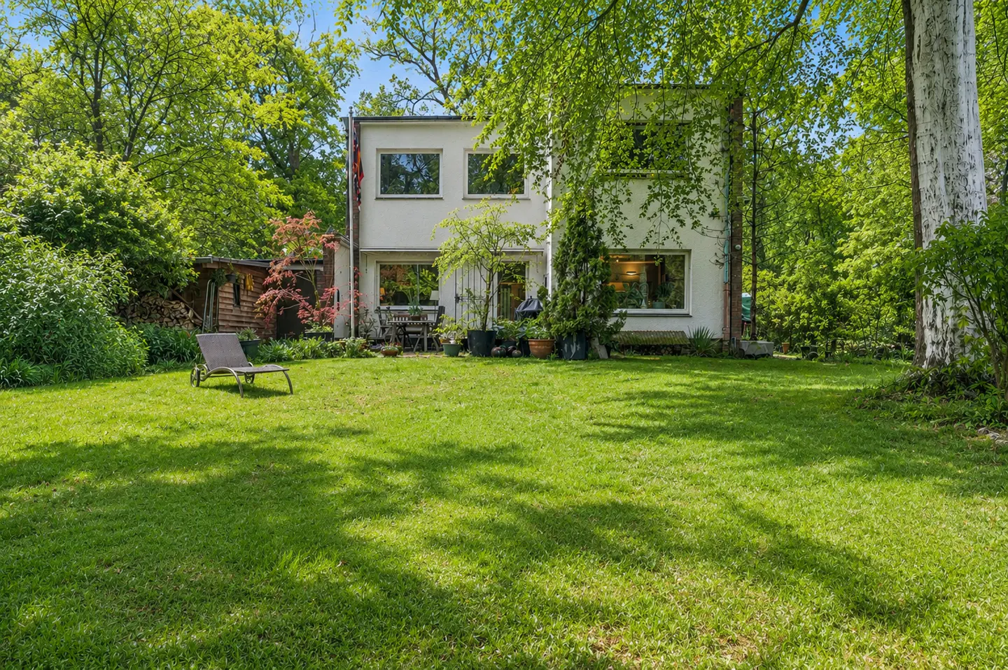 Two-story white house with a large green lawn and trees. A lounge chair sits on the grass. A table and chairs are on the patio.