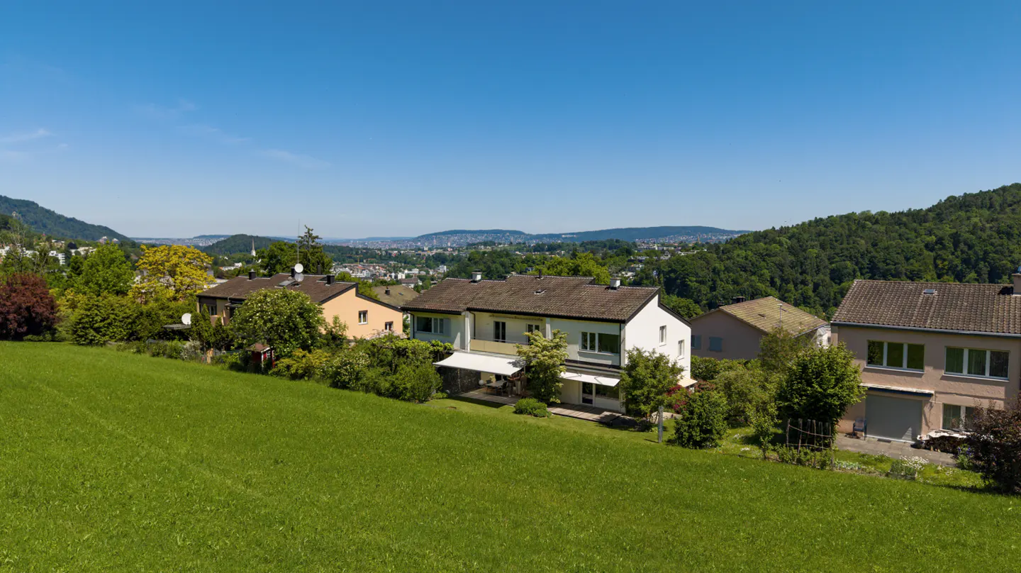 A sunny, wide shot of houses on a green hillside with a city and mountains in the distance.