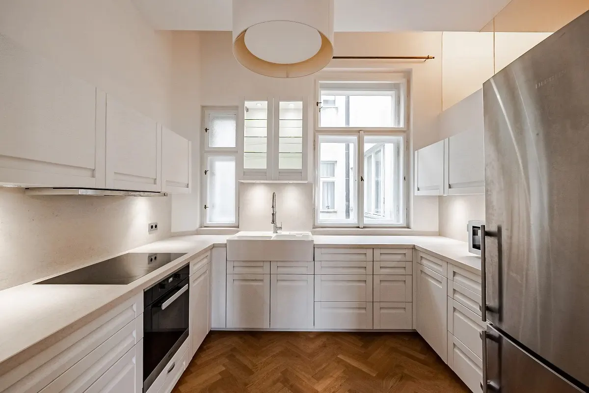 Bright kitchen with white cabinets, countertops, and a farmhouse sink under a window. Stainless steel fridge and herringbone wood floor.