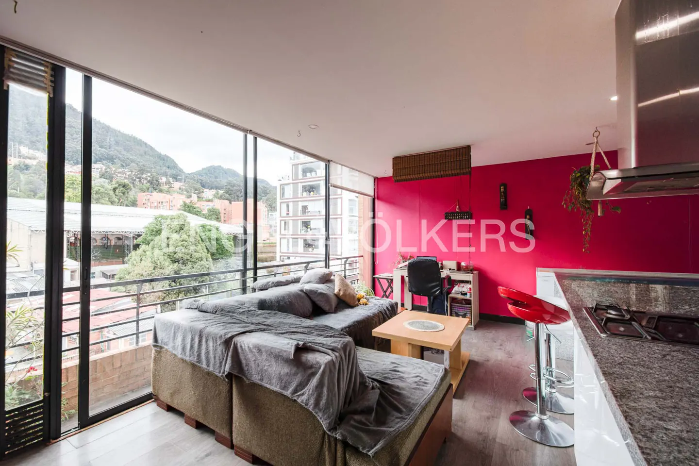 Bright apartment interior with a gray sofa, wooden table, and red accent wall. Large windows offer a city view.