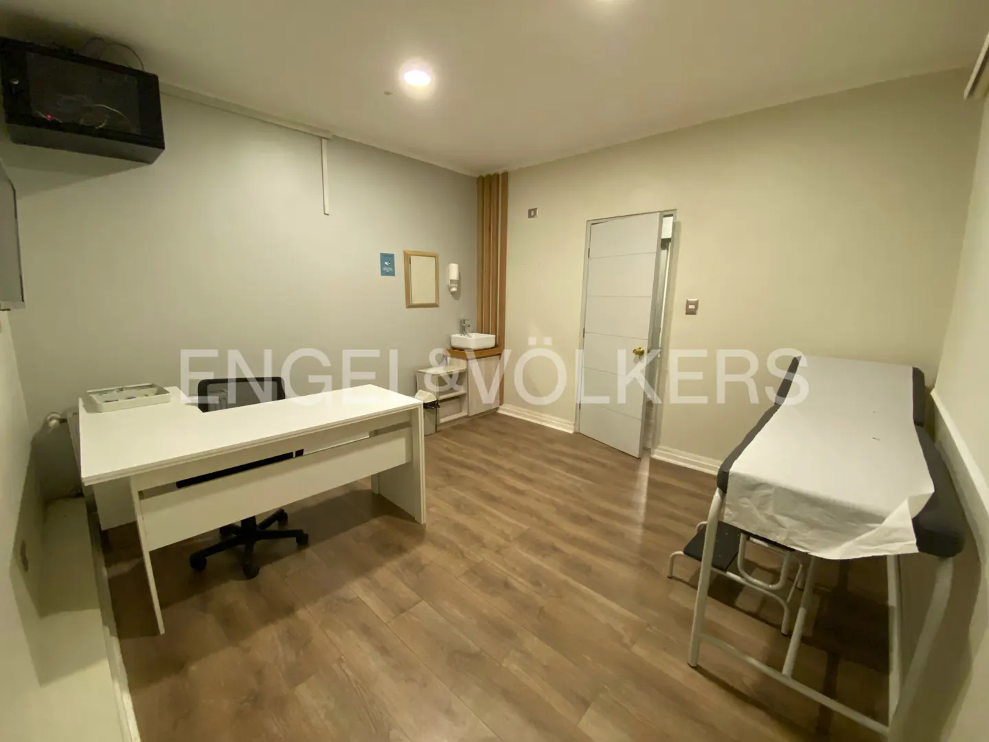 Bright medical office with a white desk, exam table, sink, and TV. The floor is wood and the walls are light gray.