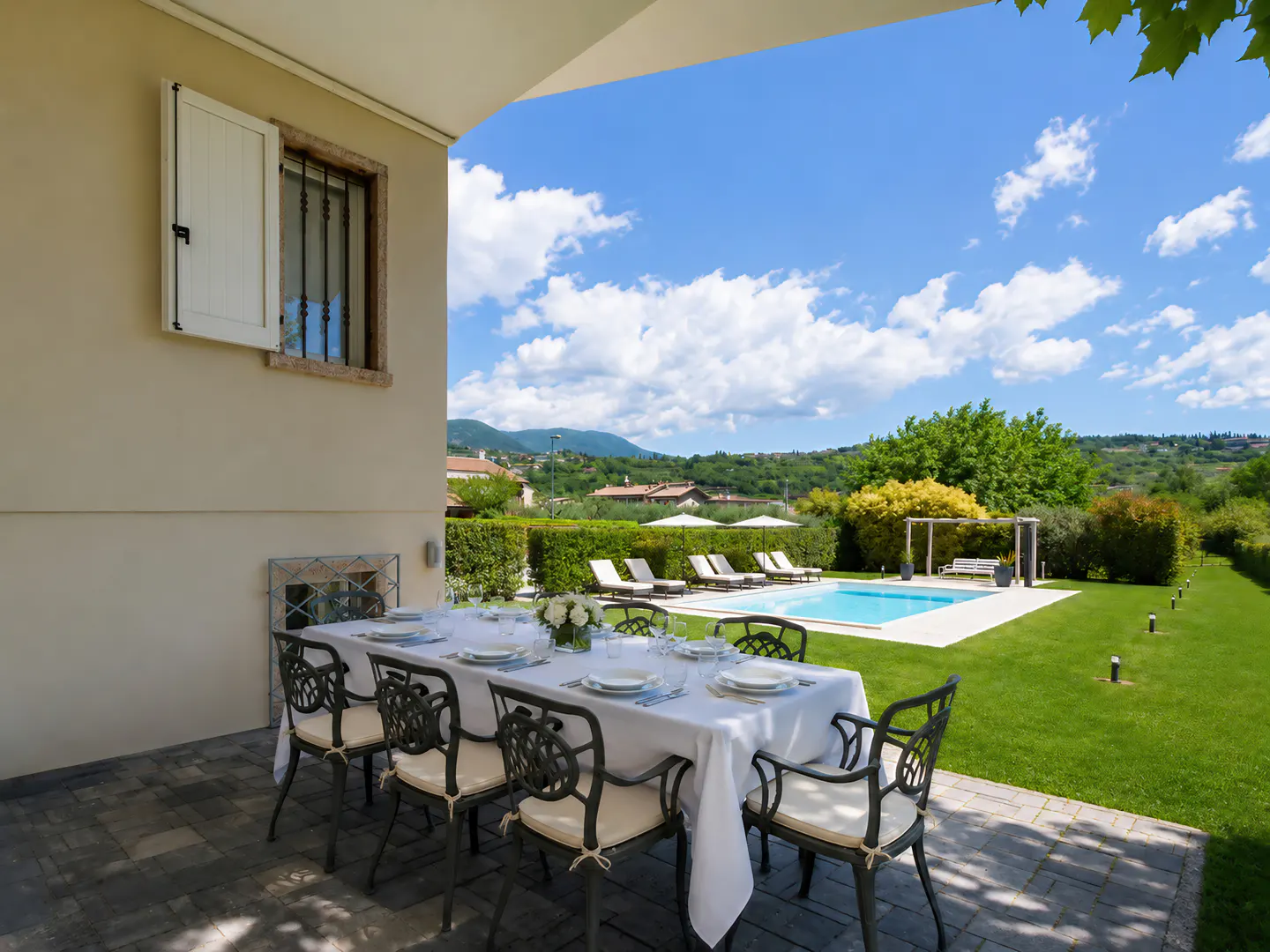 Outdoor dining table set on a patio overlooking a sunny backyard with a swimming pool, green lawn, and distant hills.