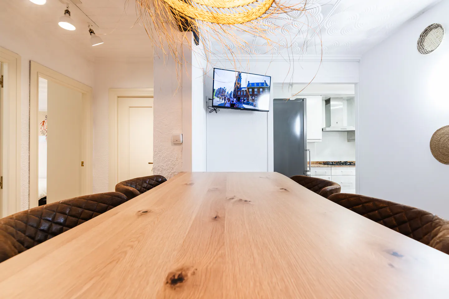 A bright dining room features a long wooden table with brown leather chairs, a TV, and a view into the kitchen.