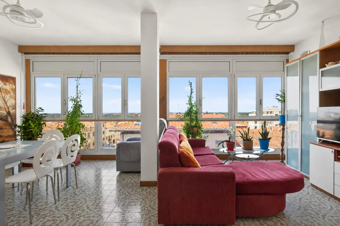 Bright living room with a red sectional sofa, dining area, patterned tile floors, and large windows overlooking a city.