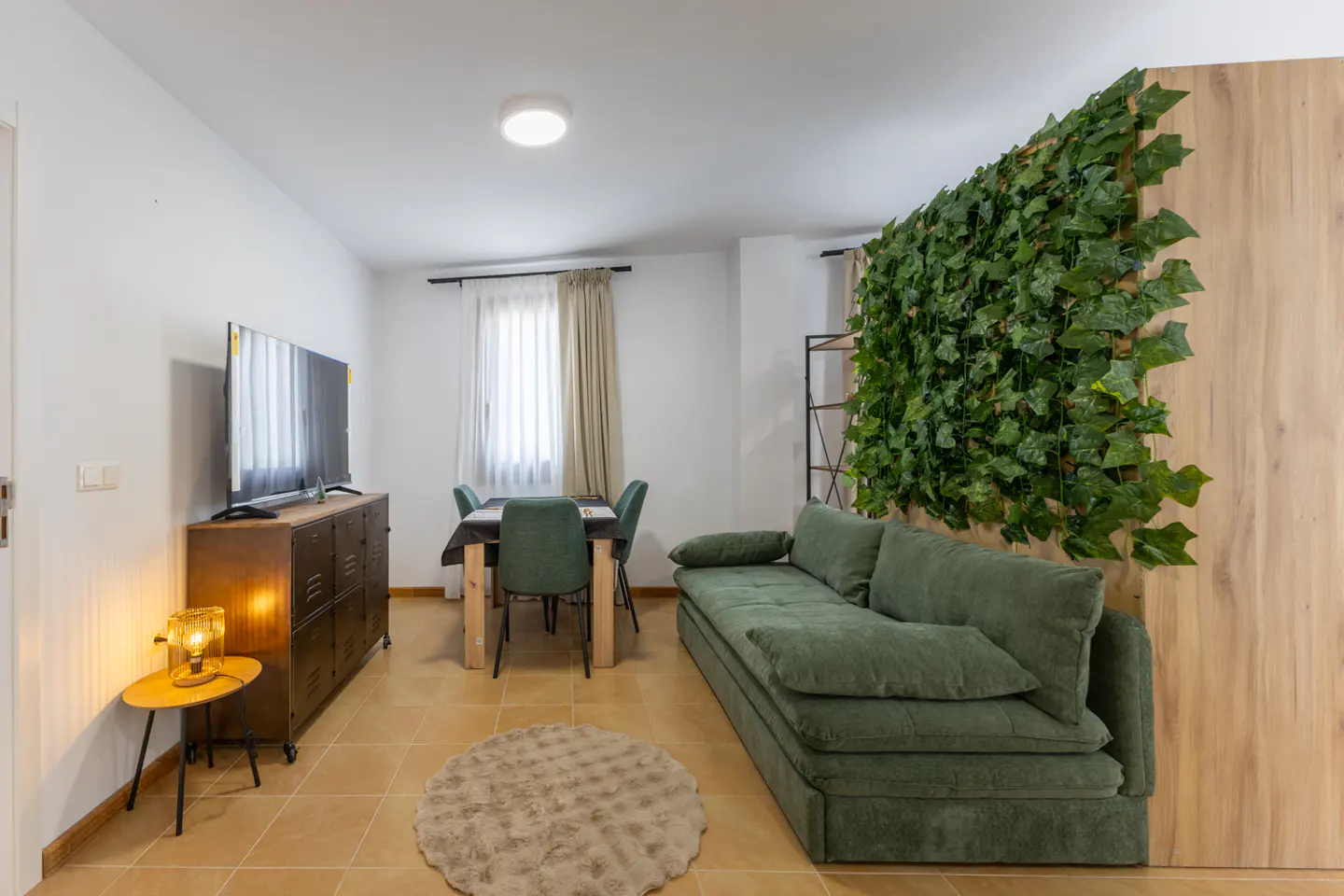 Living room with green sofa, dining table, and TV. A wall of green ivy adds a natural touch. Tiled floor and warm lighting.