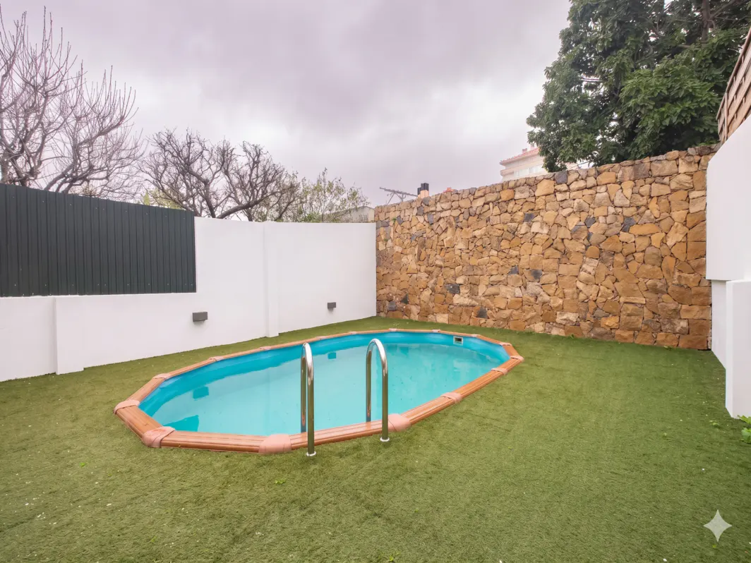 Backyard view of an in-ground pool with metal ladder, surrounded by green grass, a stone wall, and a white fence.