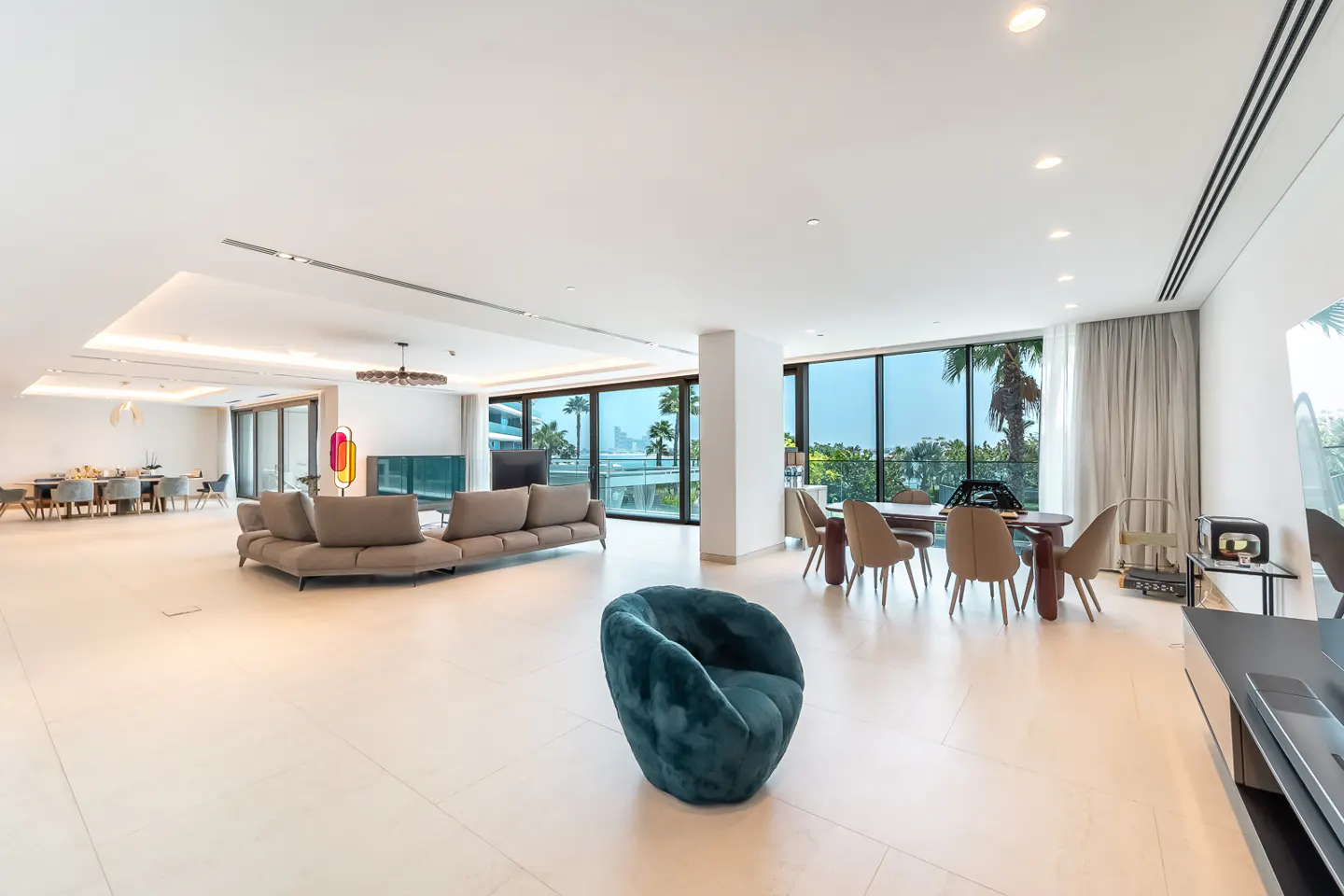 Bright, open living space with a gray sofa, teal chair, and dining table. Large windows offer a view of palm trees and the ocean.
