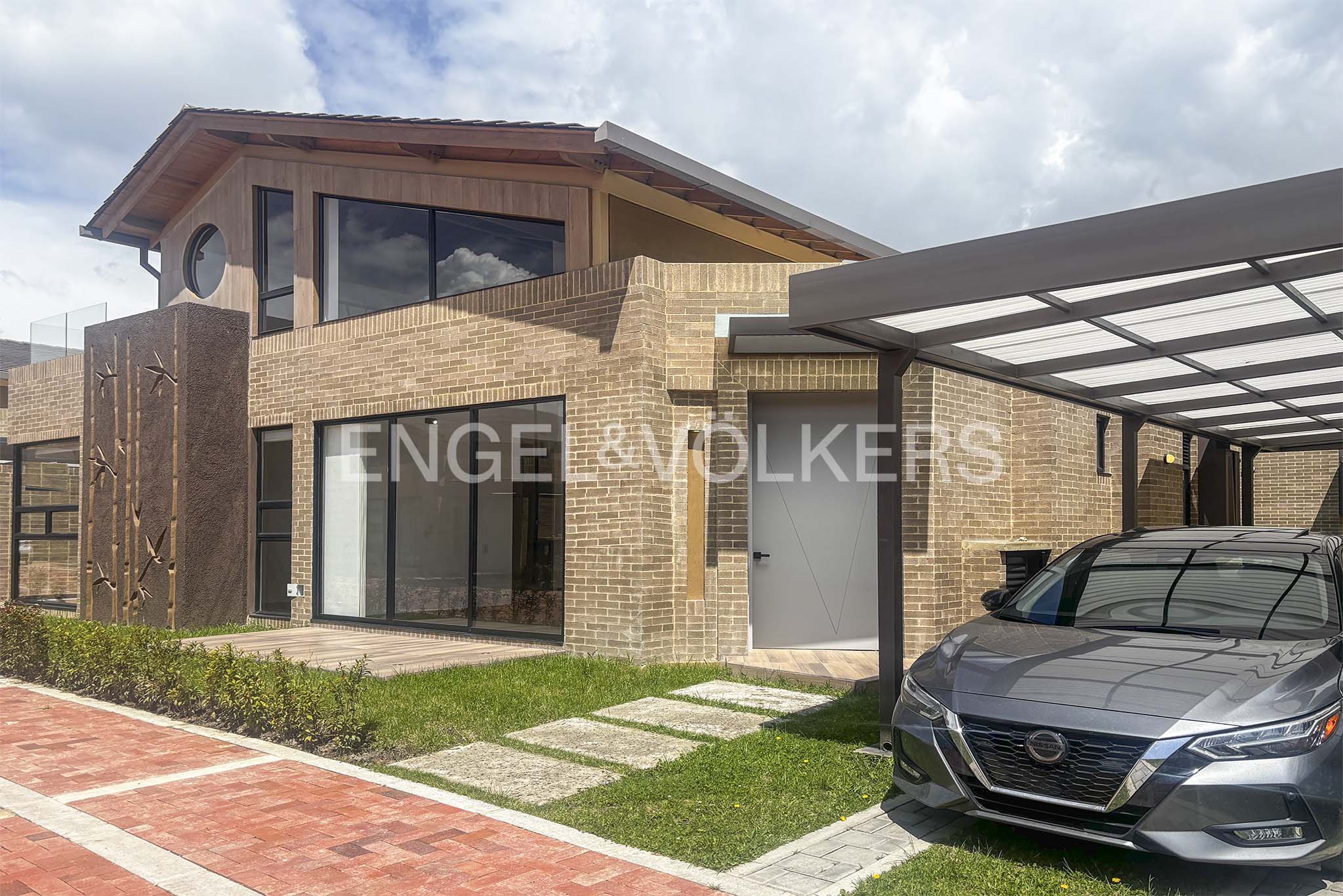 Exterior of a modern two-story brick house with a gray car parked under a carport. The sky is cloudy.