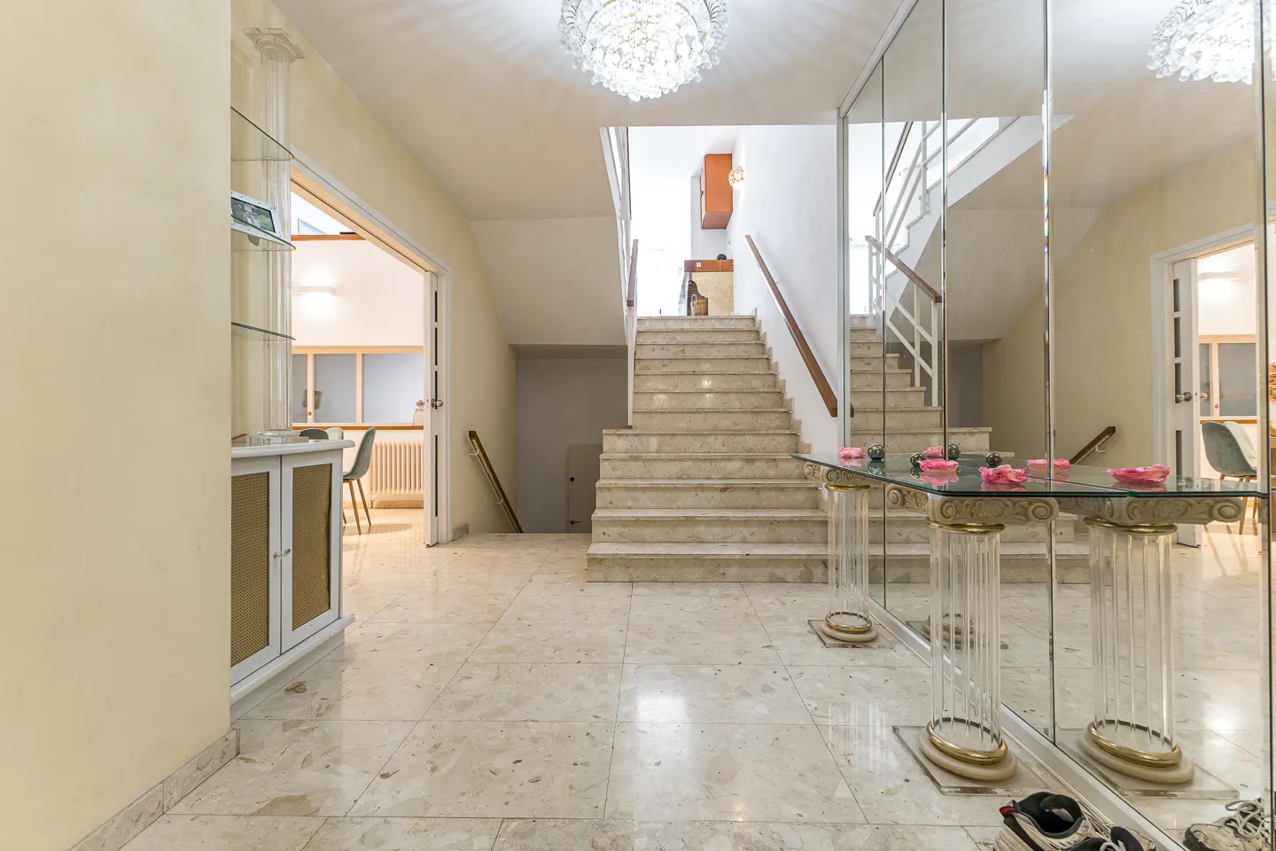 Bright foyer with marble floors, a staircase, and a mirrored wall. A glass table with pink petals sits in front of the mirror. A chandelier hangs from the ceiling.