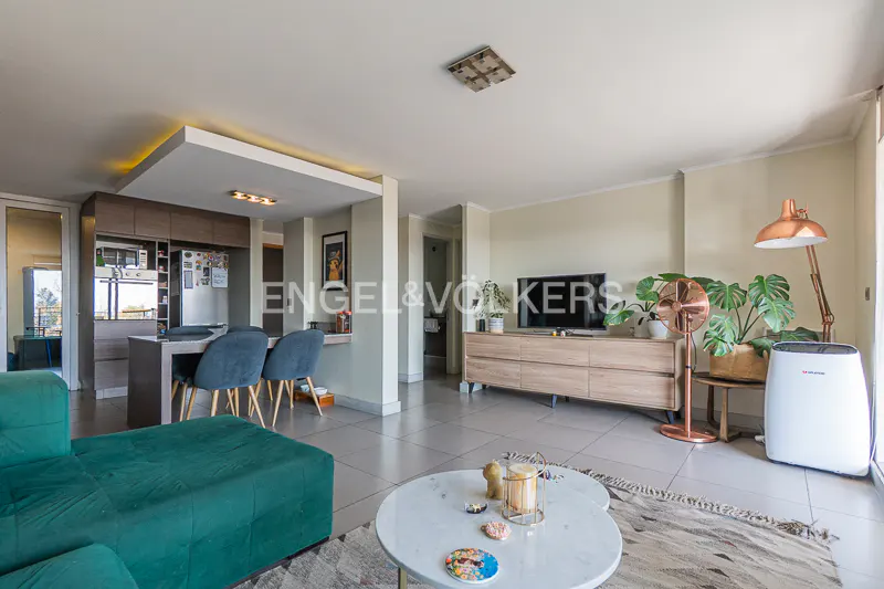 Open-concept living space with a green sofa, marble coffee table, and a kitchen with blue chairs. A TV and plants sit on a wooden console.
