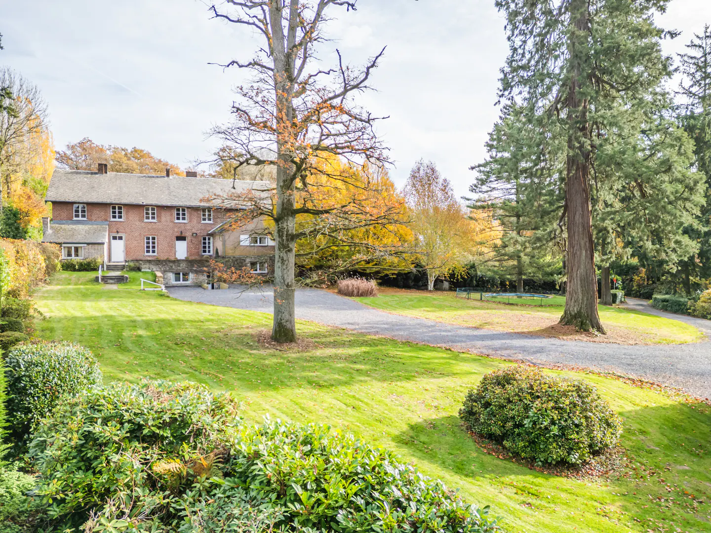 Exterior view of a two-story brick house with white trim, surrounded by green lawn, trees, and a gravel driveway.