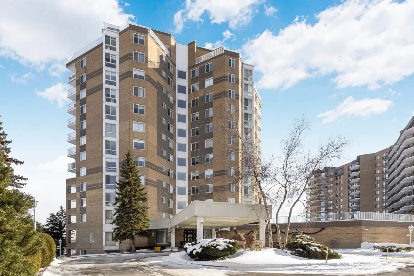 Exterior view of a tall, tan brick apartment building with white balconies and a covered entrance on a snowy day.