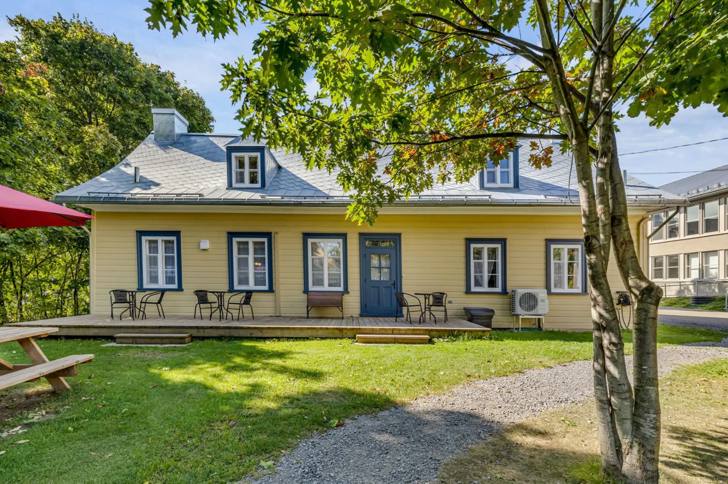 Exterior of a yellow house with blue trim and a gray roof. A wooden deck with chairs is in front. A tree is on the right.